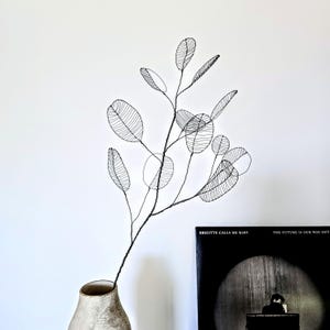 Può includere: Una scultura in filo metallico nero a forma di ramo frondoso, con linee sottili e delicate che formano foglie ovali. La scultura è posizionata in un vaso testurizzato di colore chiaro accanto a una copertina di disco nera intitolata Brigitte Calls Me Baby.