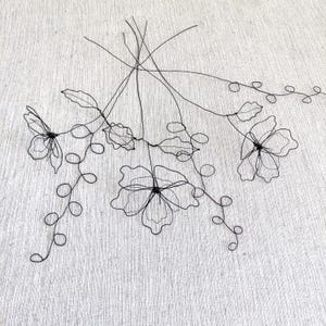 May include: A decorative sculpture made of thin black wire shaped into three distinct flowers with delicate petals and long, winding stems featuring small circular loops. The structure is composed of interconnected wire lines forming floral silhouettes.
