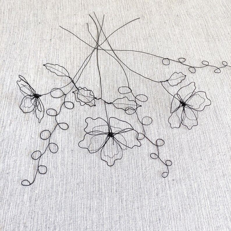 May include: A decorative sculpture made of thin black wire shaped into three distinct flowers with delicate petals and long, winding stems featuring small circular loops. The structure is composed of interconnected wire lines forming floral silhouettes.