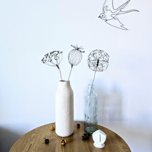 May include: A still life arrangement featuring wire sculptures of flowers in vases. The flowers include a dandelion, a seed pod, and a cluster of small blooms. A wire bird is mounted on the wall above the table. The vases are white and clear glass.