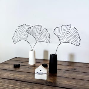 Könnte beinhalten: Zwei schwarze Drahtskulpturen von Ginkgoblättern in weißen und schwarzen Vasen auf einem Holztisch mit einem weißen hausförmigen Objekt mit dem Wort "HOME" darauf.