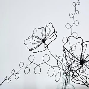 May include: A black wire sculpture shaped into two flowers with delicate, thin petals and long, winding stems featuring circular loops. The sculpture is positioned inside a clear glass bottle, creating a minimalist, linear floral arrangement.