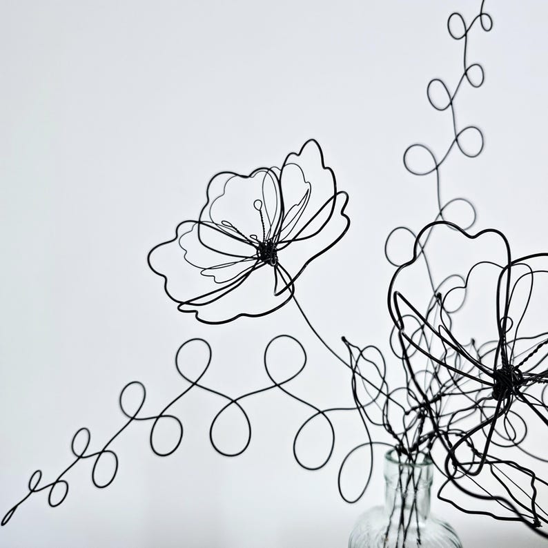 May include: A black wire sculpture shaped into two flowers with delicate, thin petals and long, winding stems featuring circular loops. The sculpture is positioned inside a clear glass bottle, creating a minimalist, linear floral arrangement.