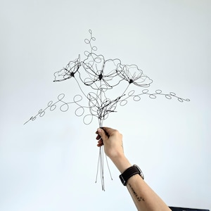 May include: A black wire sculpture shaped like a bouquet of flowers with three distinct blossoms and two long, curling leafy stems. The sculpture is constructed from thin, continuous black metal wire, forming delicate outlines of petals and foliage.