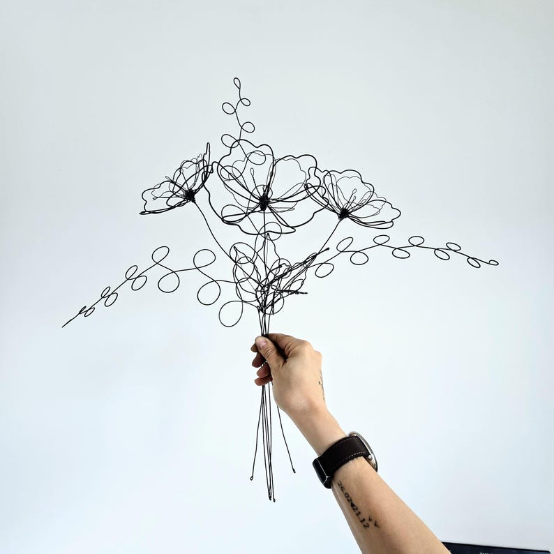 May include: A black wire sculpture shaped like a bouquet of flowers with three distinct blossoms and two long, curling leafy stems. The sculpture is constructed from thin, continuous black metal wire, forming delicate outlines of petals and foliage.