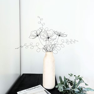 May include: A black wire sculpture shaped like a bouquet of flowers and leafy stems, featuring thin, continuous metal lines forming floral outlines and swirling foliage. The sculpture is displayed inside a tall, cylindrical, off-white vase.