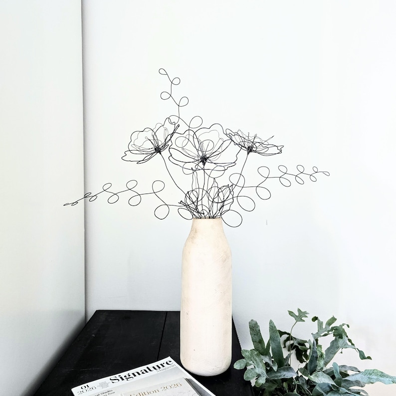 May include: A black wire sculpture shaped like a bouquet of flowers and leafy stems, featuring thin, continuous metal lines forming floral outlines and swirling foliage. The sculpture is displayed inside a tall, cylindrical, off-white vase.