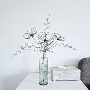 May include: A black wire sculpture shaped like a bouquet of three flowers with swirling stems and leaves, placed inside a clear, vertically ribbed glass bottle. The wire art features intricate loops and lines forming floral silhouettes.