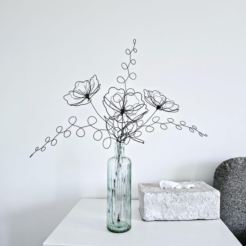 May include: A black wire sculpture shaped like a bouquet of three flowers with swirling stems and leaves, placed inside a clear, vertically ribbed glass bottle. The wire art features intricate loops and lines forming floral silhouettes.