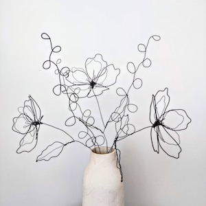 May include: A black wire sculpture shaped into a bouquet of three flowers with swirling stems and leaves. The sculpture is placed inside a textured, off-white ceramic vase. The wire forms delicate, open outlines of petals and foliage.
