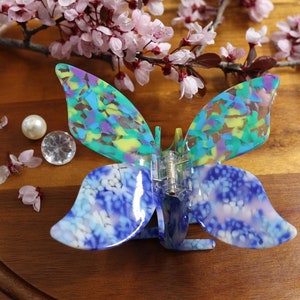 May include: A blue and green butterfly-shaped hair clip with a marbled pattern. The clip is on a wooden surface with pink flower petals.