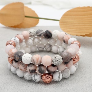 May include: Three layered bracelets with white, pink, and gray beads. The beads are made of lava rock, white howlite, and rose quartz. The bracelets have rose gold accents.