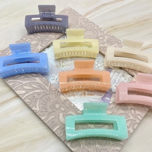May include: A collection of rectangular hair claw clips in various pastel colors, including blue, yellow, peach, pink, and green. The clips are made of a translucent material and are arranged on a patterned surface.