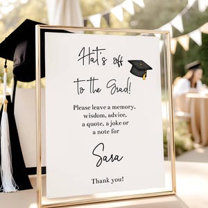 May include: A graduation sign in a gold frame with the text "Hat's off to the Grad!" and "Sara". The sign includes a graduation cap illustration. The background is blurred with outdoor elements.