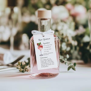Puede incluir: Una pequeña botella de vidrio transparente llena de líquido rosa, con una tapa de madera clara. Una etiqueta blanca atada indica "Rose Spritzer" y detalla una receta de cóctel. El fondo está borroso, con flores.