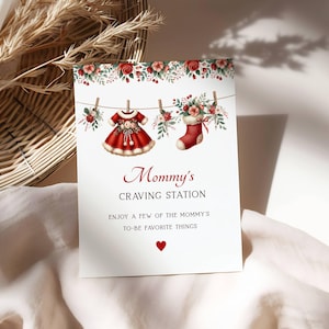Könnte beinhalten: Eine weiße Karte mit dem Text "Mommy's Craving Station" in burgunderfarbener Schrift, mit einem roten Babykleid und Strumpf, die an einer Wäscheleine hängen. Blumige Akzente und ein Herz vervollständigen das Design, ideal für eine Babyparty.