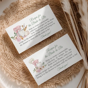 May include: Two recipe cards for a bridal shower, featuring text and watercolor illustrations. The cards read "Recipes for the Bride-to-Be" and include instructions for guests to share recipes. The cards are on a woven placemat.