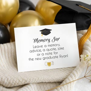 May include: A white card with the text "Memory Jar" and instructions to leave a note for the new graduate Ryan. The card is next to a graduation cap, gold and black balloons, and a white knitted blanket.