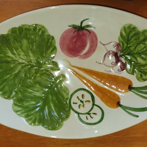 May include: A white ceramic serving platter with a painted design of vegetables. The platter features a green leafy vegetable, a red tomato, a white radish, two orange carrots, and two green cucumber slices.