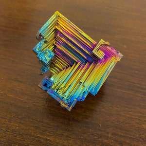 May include: A colorful bismuth crystal with a geometric, stepped structure. The crystal displays a vibrant array of colors, including blues, yellows, purples, and greens. The surface has a metallic sheen, and the crystal is set against a dark wooden background.