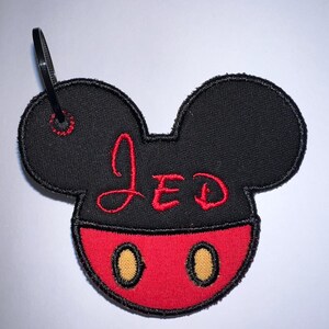 May include: A black and red embroidered patch shaped like a mouse head with rounded ears. The top section is black with the name JED stitched in red thread. The bottom section is red with two yellow oval accents. A black plastic zip tie is attached.