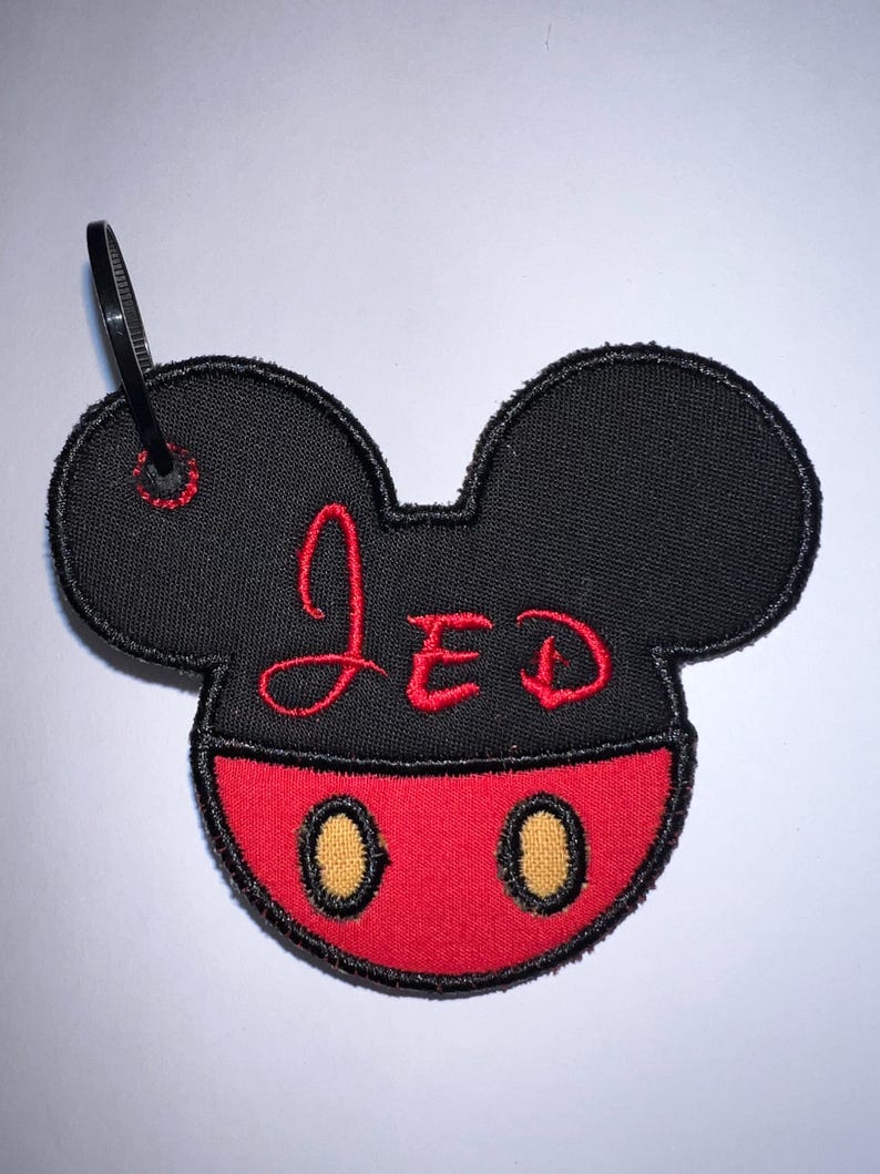 May include: A black and red embroidered patch shaped like a mouse head with rounded ears. The top section is black with the name JED stitched in red thread. The bottom section is red with two yellow oval accents. A black plastic zip tie is attached.