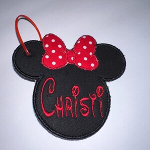 May include: A black fabric ornament shaped like a mouse head with a red and white polka dot bow. The name Christi is embroidered in red thread across the center. A thin red plastic loop is attached to the top left ear for hanging purposes.