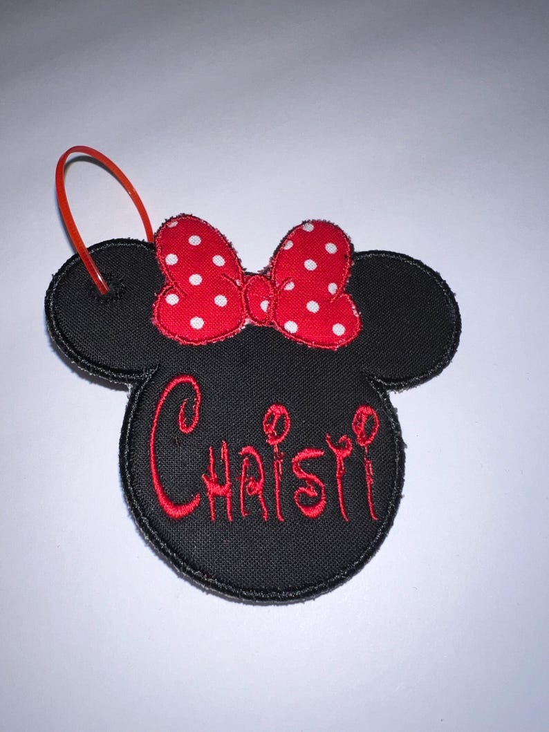 May include: A black fabric ornament shaped like a mouse head with a red and white polka dot bow. The name Christi is embroidered in red thread across the center. A thin red plastic loop is attached to the top left ear for hanging purposes.