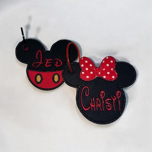 May include: Two embroidered fabric keychains shaped like mouse heads. One is black with red shorts, yellow buttons, and the name JED in red thread. The other is black with a red and white polka dot bow and the name CHRISTI in red thread.