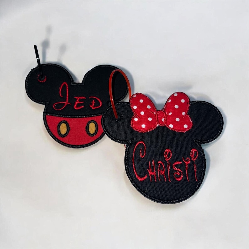 May include: Two embroidered fabric keychains shaped like mouse heads. One is black with red shorts, yellow buttons, and the name JED in red thread. The other is black with a red and white polka dot bow and the name CHRISTI in red thread.