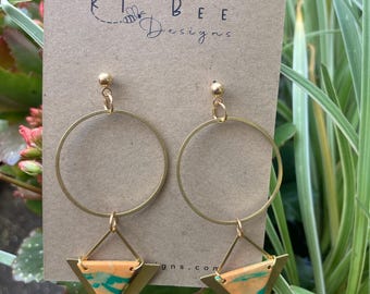 May include: A pair of gold-colored hoop earrings with a geometric design. Each earring features a large circular hoop connected to a brass triangle with orange and teal accents. The earrings are displayed on a brown card with the text "KT Bee Designs".
