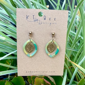 May include: A pair of dangle earrings with a leaf design. The earrings feature a gold-colored leaf charm inside a teardrop-shaped frame with shades of green and yellow. The earrings are displayed on a brown card with the text "KT Bee Designs" and "ktbeedesigns.com".