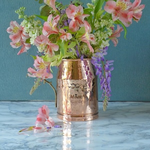Puede incluir: Un ramo de flores de alstroemeria rosas y blancas, lilas blancas y glicinias moradas en un jarrón de cobre martillado con la inscripción "Jardín de mamá".
