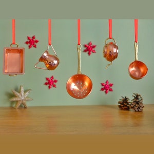 May include: Five miniature copper kitchen utensils hanging from red string. The utensils include a small pot, a pitcher, a strainer, a ladle, and a tea kettle. The utensils are hanging against a green background with red snowflake decorations.