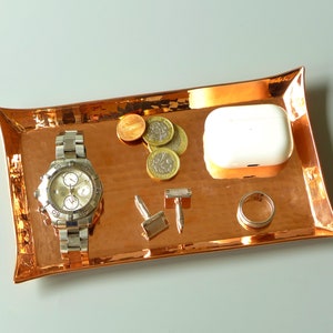 Puede incluir: Una bandeja de cobre con un acabado martillado que contiene un reloj plateado, gemelos, un anillo y una funda de carga inalámbrica blanca. También hay varias monedas de oro en la bandeja.