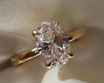 Puede incluir: Un anillo de compromiso de oro con un diamante de talla ovalada. El diamante está engastado con cuatro garras y la banda es de oro pulido. El anillo se muestra sobre una tela de satén blanca.