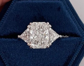 Può includere: Una scatola per anelli esagonale con un interno in velluto blu. Un anello di fidanzamento con diamante con una grande pietra centrale di forma quadrata e due pietre triangolari più piccole su entrambi i lati è esposto nella scatola.