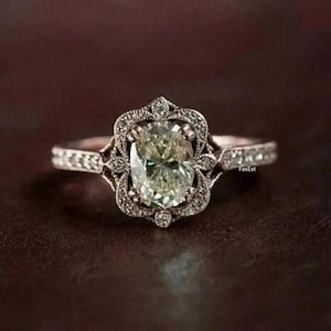 Puede incluir: Un anillo de plata con un diamante de forma ovalada engastado en un diseño delicado y ornamentado. El anillo presenta un patrón de filigrana con pequeños diamantes que rodean la piedra central.