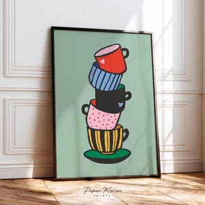 May include: A framed art print depicting a stack of vibrant teacups. The cups are red, blue, black, pink, and yellow, adorned with patterns like stripes, polka dots, and hearts. The background is a pale green. The print is signed "Paper Melon Prints".