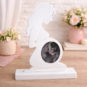 Op de afbeelding: Witte houten silhouet van een zwangere vrouw, met een cirkelvormig frame met een zwart-wit echografiebeeld. Het frame staat op een rechthoekige basis. Bloemen in een roze vaas op de achtergrond.