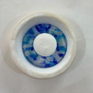 May include: A circular white ceramic bowl featuring a central white raised disc surrounded by a ring of embedded blue and turquoise glass fragments. The interior base displays a mosaic-like pattern of translucent coloured glass pieces.