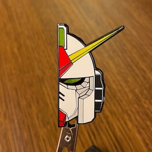 Könnte beinhalten: Eine weiße, rote, grüne und gelbe Emaille-Anstecknadel, die den Kopf eines Roboters darstellt. Das Auge des Roboters ist grün und die Nadel ist an einem silbernen Metallverschluss befestigt.