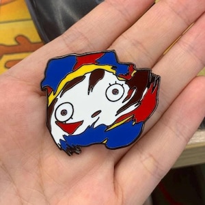 May include: A metal enamel pin featuring a stylised, abstract face with a white base, large circular eyes, and a red mouth. The design is framed by bold, irregular shapes in primary colours including deep blue, bright red, and yellow, with dark brown accents.