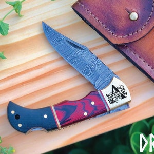 Könnte beinhalten: Ein personalisiertes Taschenmesser mit einer Damastmesserklinge und einem rot-schwarzen Holzgriff. Das Messer ist mit dem Buchstaben "A" und dem Namen "Anderson" graviert. Es wird mit einer braunen Lederscheide geliefert.