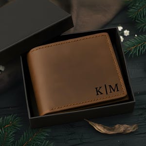 May include: A brown leather wallet with the initials "K|M" embossed on the front, presented in a black gift box. The wallet is a bi-fold style, with visible stitching around the edges. The background includes greenery and a dark surface.