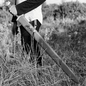 May include: A person holding a black and silver sword with a leather scabbard. The sword has a decorative design on the blade and a cross-shaped guard.