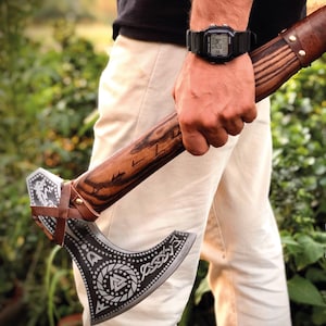 May include: A hand holding a Viking axe with a wooden handle and a leather strap. The axe head is decorated with a silver design and a dragon. The text "NORSE OLAF" is visible in the background.