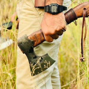 May include: A person holding a black and brown axe with a leather handle and a brown leather strap. The axe has a dark metal blade and a wooden handle. The person is wearing a beige shirt and a black watch with a digital display.
