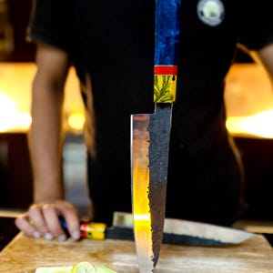 Pu&ograve; includere: Un primo piano di un coltello da chef con una lama nera e argentata e un manico colorato. Il coltello &egrave; in piedi su un tagliere di legno con cipolle e lime tagliate a fette.
