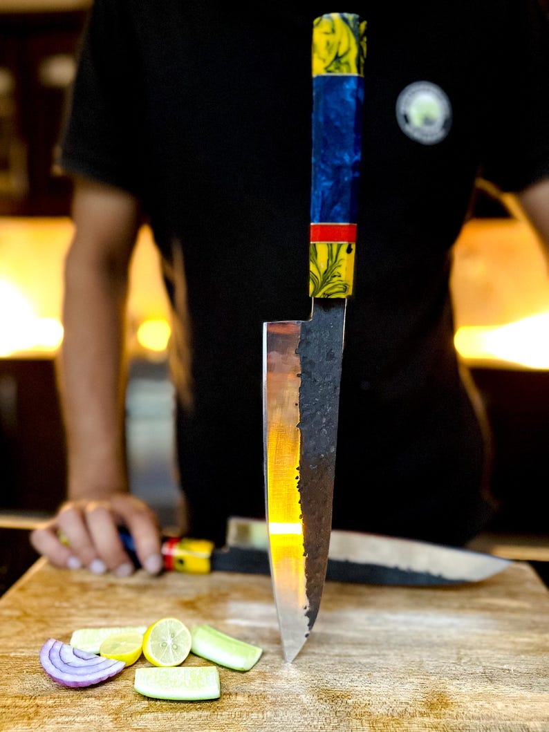 Pu&ograve; includere: Un primo piano di un coltello da chef con una lama nera e argentata e un manico colorato. Il coltello &egrave; in piedi su un tagliere di legno con cipolle e lime tagliate a fette.